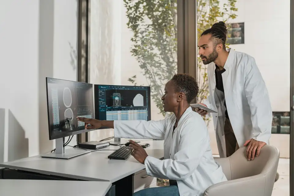 A young trainee in a white coat watching over a physician's shoulder as she points out a brain scan on a monitor